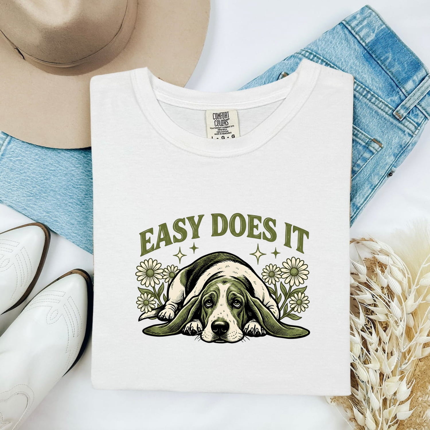 White Comfort Colors 1717 t-shirt with a basset hound graphic and 'Easy Does It' text, folded atop jeans and boots.
