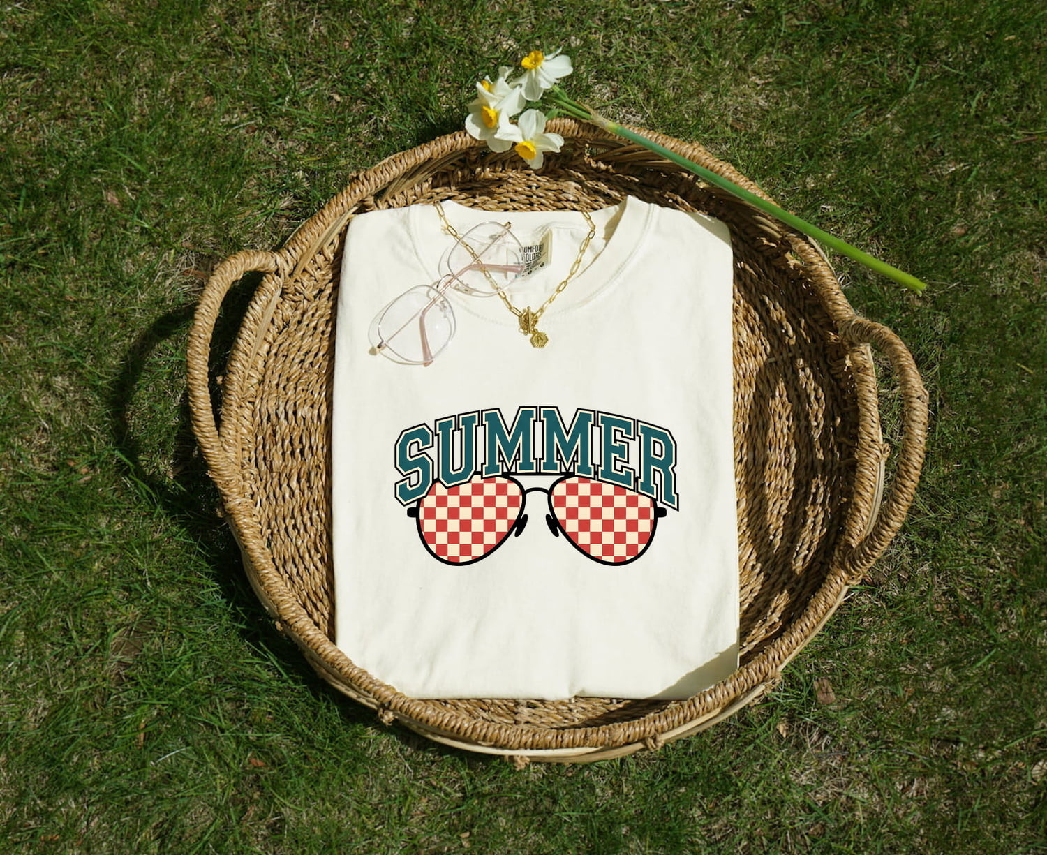 White Comfort Colors 1717 t-shirt with 'Summer' and sunglasses design folded in a woven basket on grass.