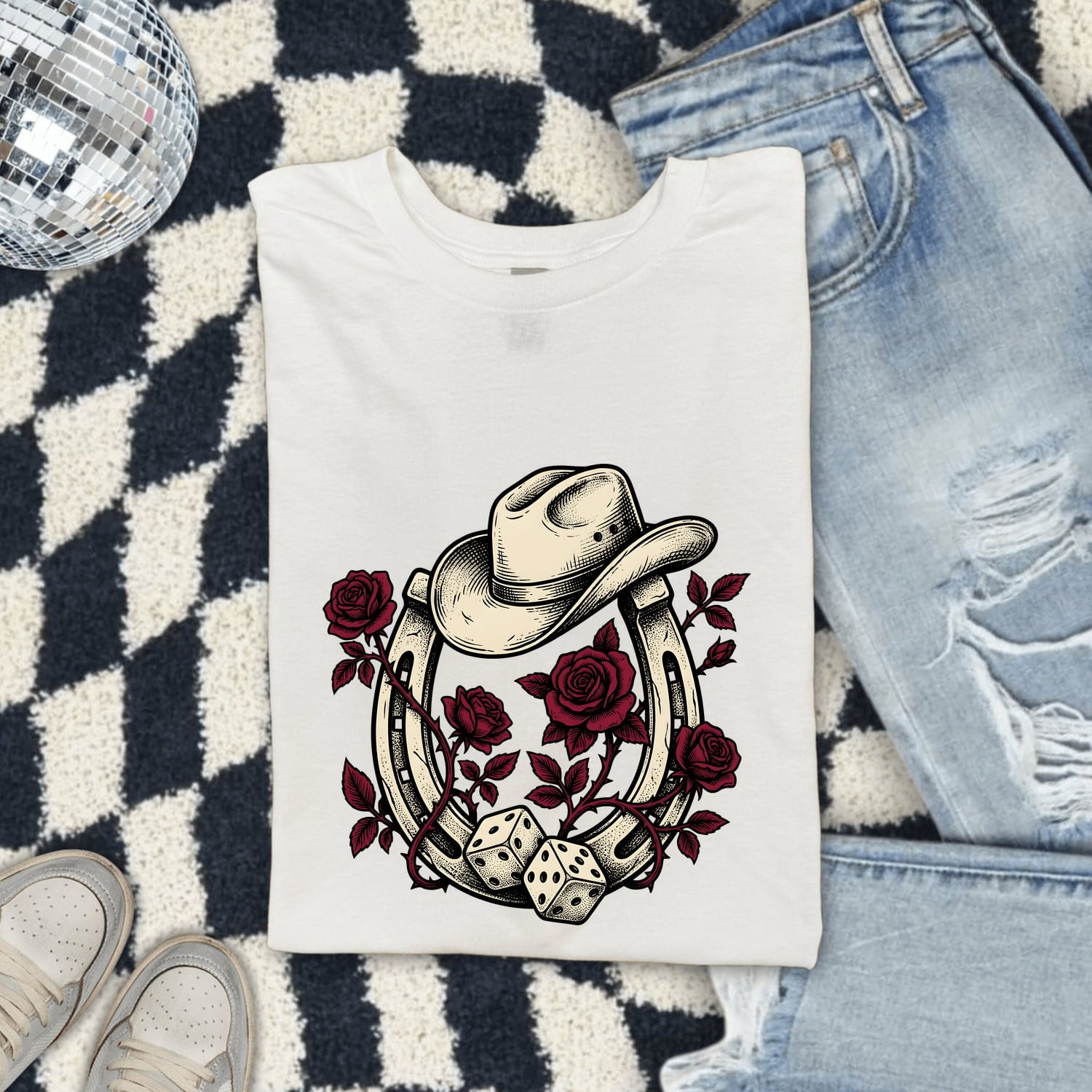 White Comfort Colors 1717 t-shirt with a cowboy hat, horseshoe, roses, and dice design, folded on a checkered rug with jeans and shoes.