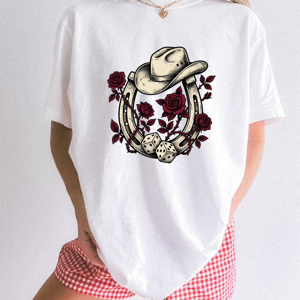 Woman wearing a white Comfort Colors 1717 t-shirt with a cowboy hat, horseshoe, roses, and dice design standing in front of a white background.