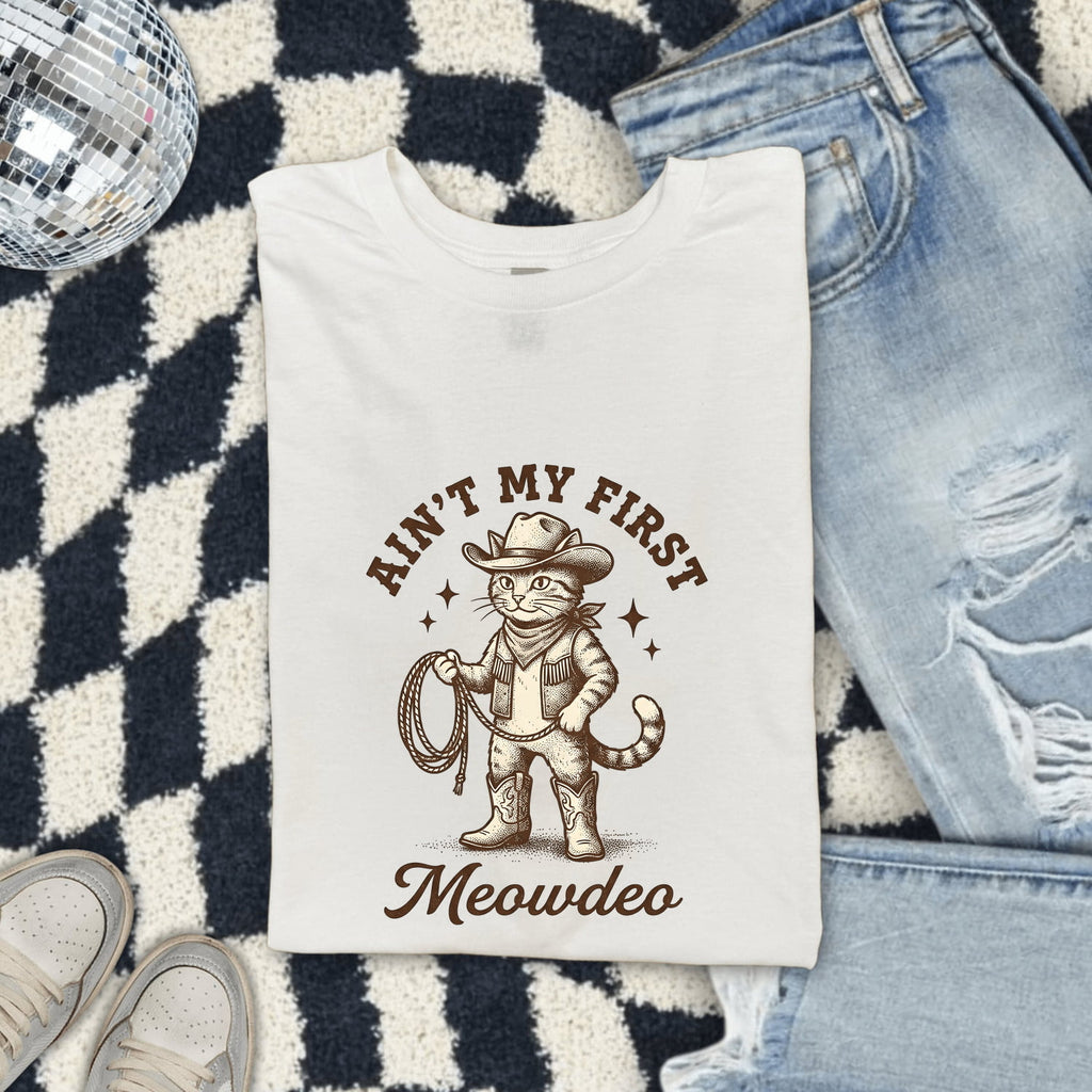 White Comfort Colors 1717 t-shirt with a cat in a cowboy outfit graphic and text, folded on a checkered rug with jeans and shoes.