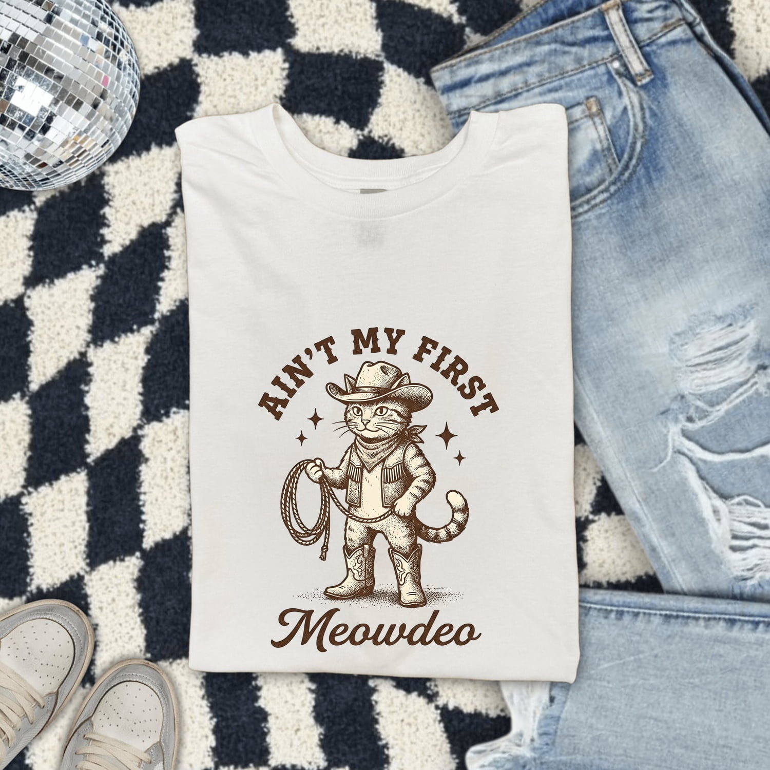 White Comfort Colors 1717 t-shirt with a cat in a cowboy outfit graphic and text, folded on a checkered rug with jeans and shoes.