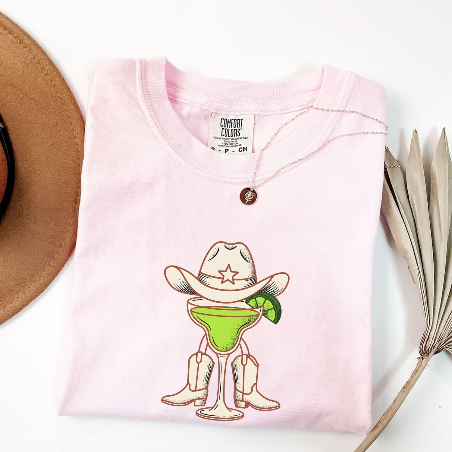Blossom Comfort Colors 1717 t-shirt with a graphic design of a margarita in cowboy boots and a hat, placed on a white surface with a hat and dried plant leaves.