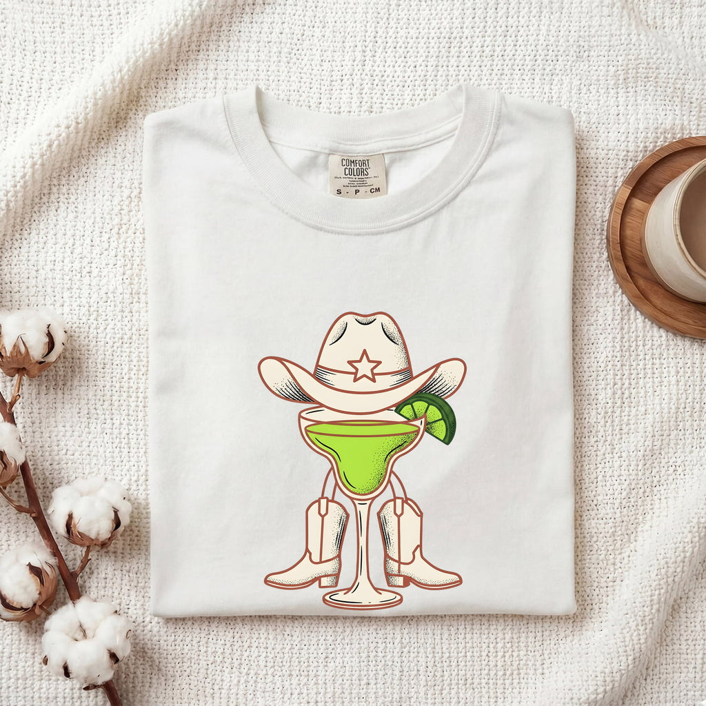 White Comfort Colors 1717 t-shirt with a graphic design of a cowboy-themed cocktail folded on a textured surface.