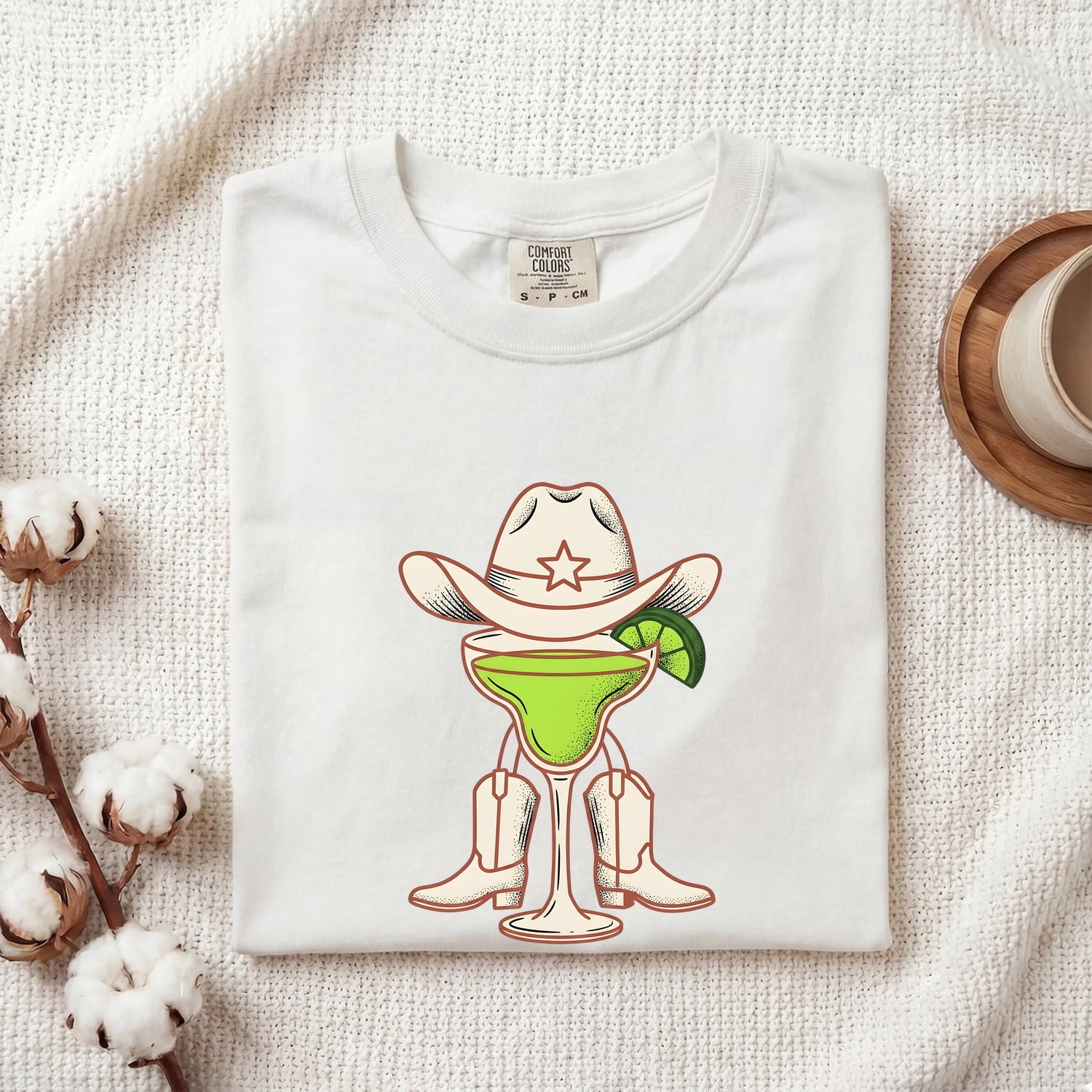 White Comfort Colors 1717 t-shirt with a graphic design of a cowboy-themed cocktail folded on a textured surface.