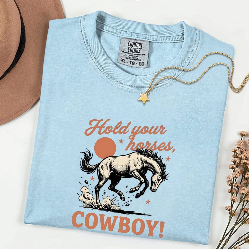 Chambray Comfort Colors 1717 t-shirt with bucking horse graphic and text 'Hold your horses, cowboy!' folded on a white background with a necklace.