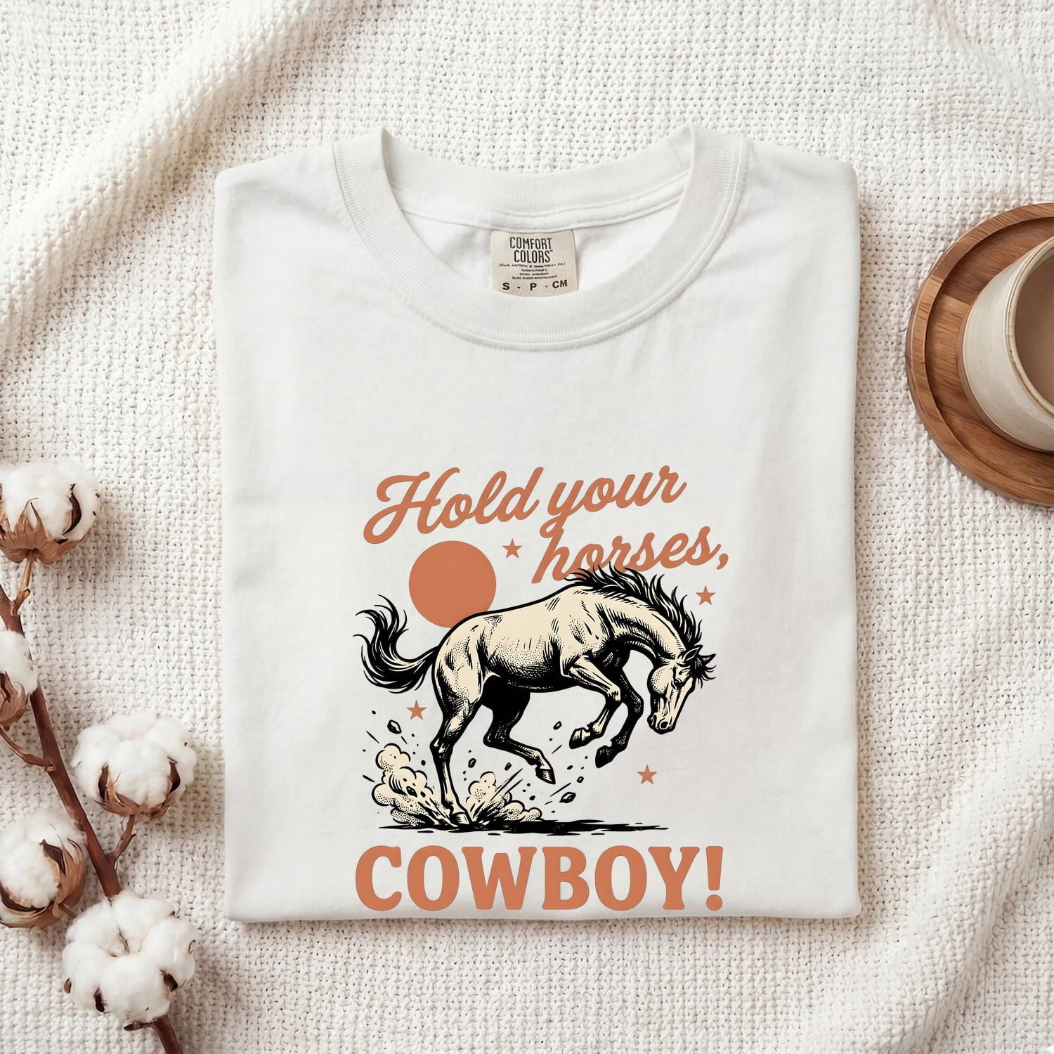 White Comfort Colors t-shirt with a bucking horse graphic and text 'Hold your horses, cowboy!', folded on a textured surface.