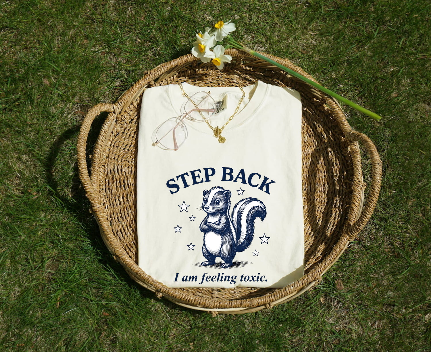White Comfort Colors 1717 t-shirt with a skunk graphic and funny text, folded in a woven basket with flowers on grass