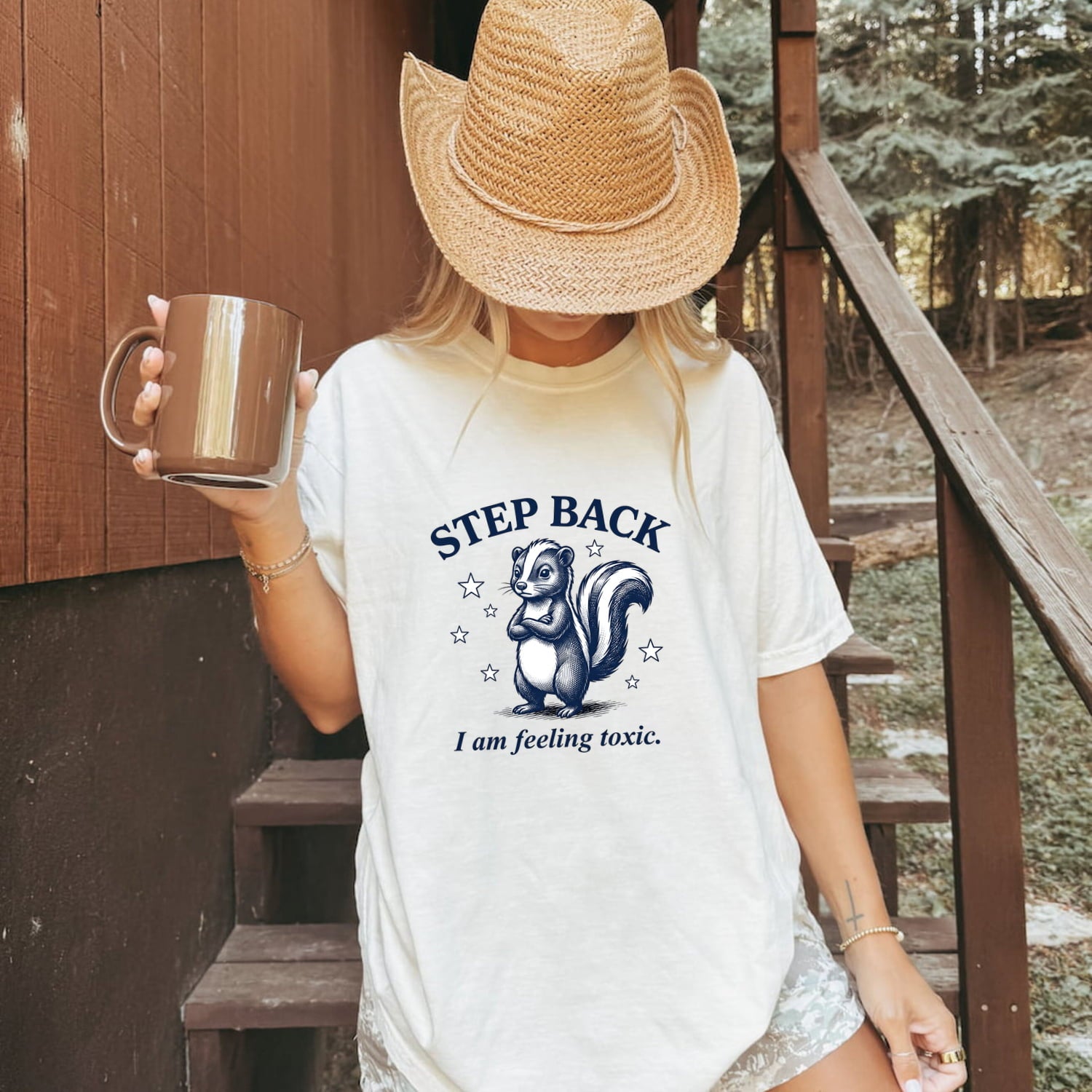Woman wearing a white Comfort Colors 1717 t-shirt with a skunk graphic and funny text, holding a mug on stairs