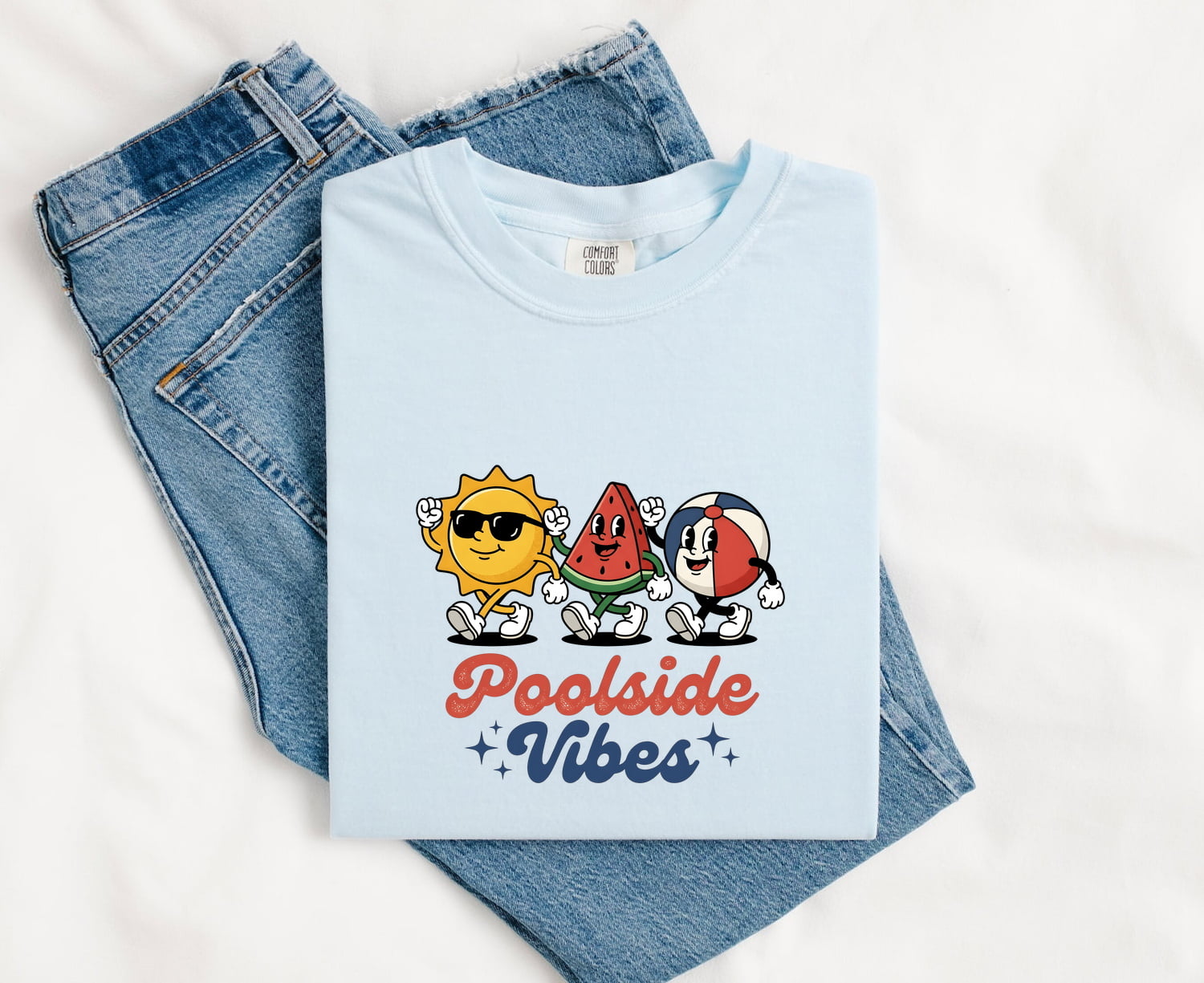 Chambray Comfort Colors 1717 t-shirt with 'Poolside Vibes' text and cartoon design, folded on blue jeans.