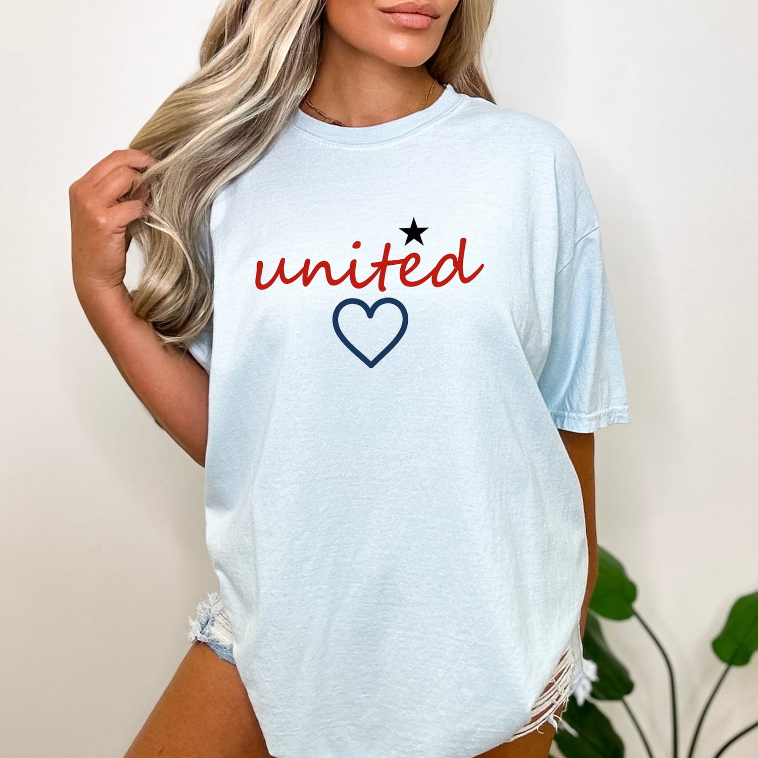 Chambray Comfort Colors 1717 t-shirt with 'united' text and heart design worn by a woman standing near a plant.