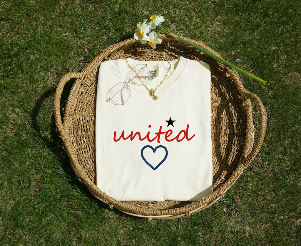 White Comfort Colors 1717 t-shirt with 'united' design, folded in a woven basket on grass